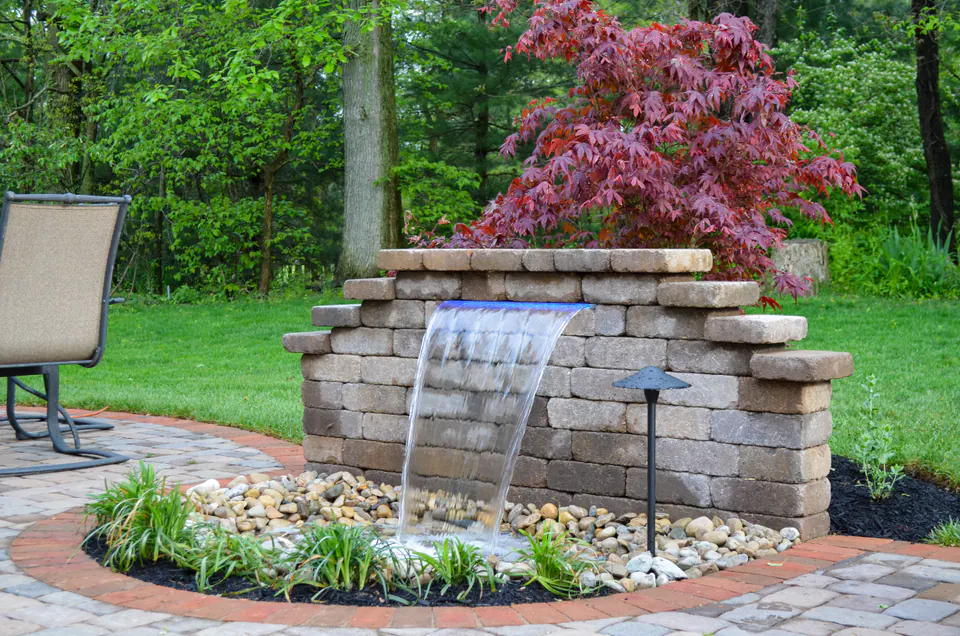 Stacked wall block waterfall with Japanese maple, river rock basin, paver patio border, and path light